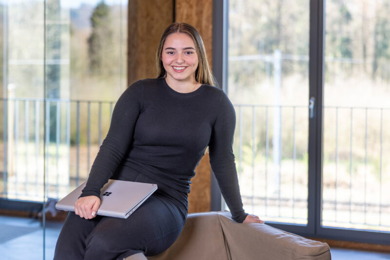 Junge Frau in schwarzem Outfit sitzt auf einer Rückenlehne, hält ein geschlossenes Microsoft Surface Laptop und lächelt in die Kamera. Im Hintergrund Fenster mit Blick ins Grüne.