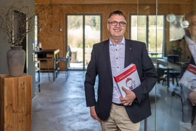 Älterer Mann im Anzug steht in einem modernen Büro und hält ein Buch mit dem Titel „Moderne Hauswartung“ in der Hand.