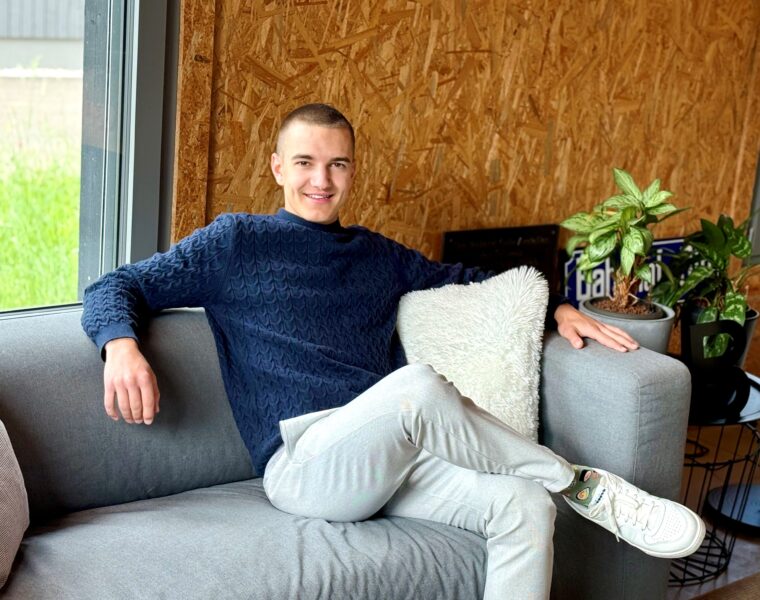 Junger Mann mit kurzem Haarschnitt sitzt entspannt auf einem grauen Sofa, trägt einen dunkelblauen Pullover und helle Hosen. Neben ihm ein weisses Kissen und grüne Zimmerpflanzen. Im Hintergrund eine Wand aus Holzplatten und ein Fenster mit Blick ins Grüne.