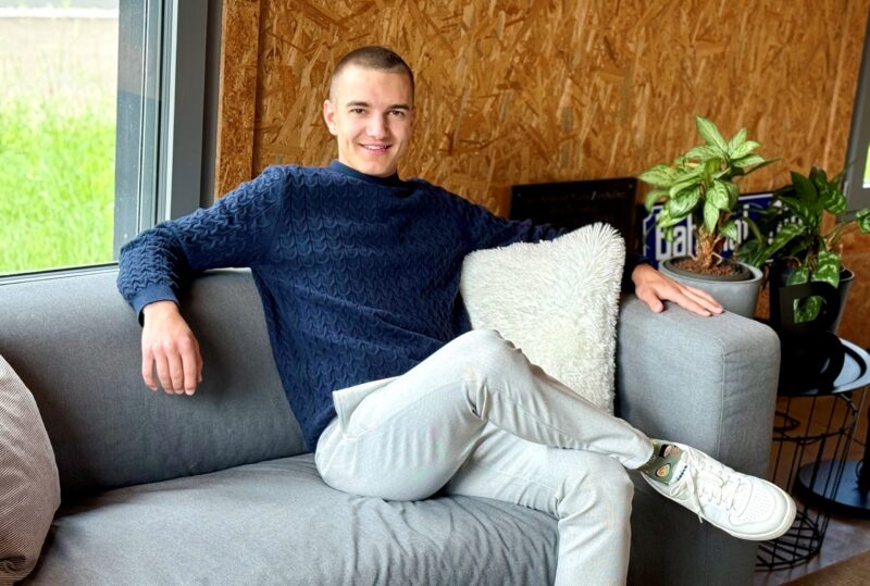 Junger Mann mit kurzem Haarschnitt sitzt entspannt auf einem grauen Sofa, trägt einen dunkelblauen Pullover und helle Hosen. Neben ihm ein weisses Kissen und grüne Zimmerpflanzen. Im Hintergrund eine Wand aus Holzplatten und ein Fenster mit Blick ins Grüne.