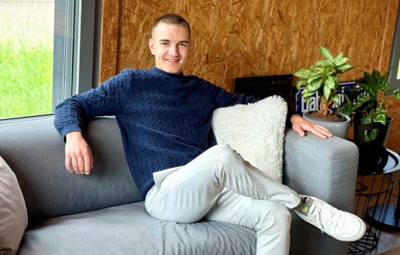 Junger Mann mit kurzem Haarschnitt sitzt entspannt auf einem grauen Sofa, trägt einen dunkelblauen Pullover und helle Hosen. Neben ihm ein weisses Kissen und grüne Zimmerpflanzen. Im Hintergrund eine Wand aus Holzplatten und ein Fenster mit Blick ins Grüne.